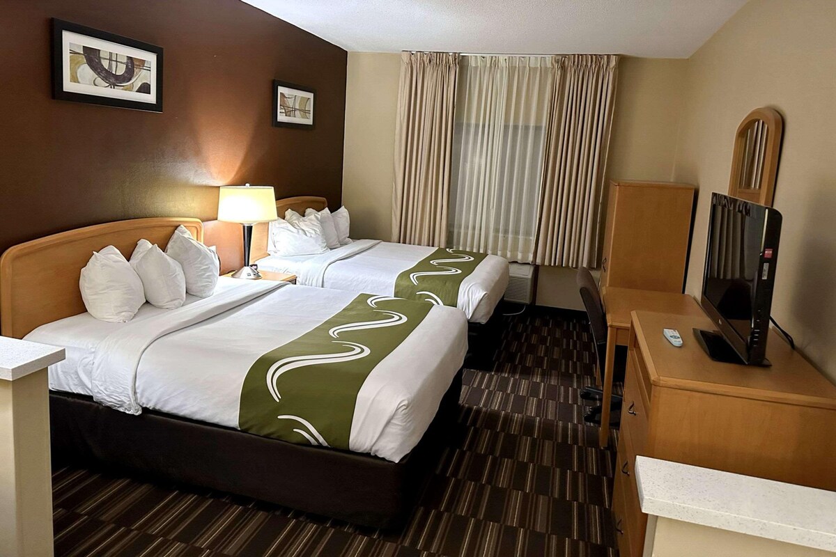 Una habitación de hotel con dos camas, un escritorio con televisor, una lámpara y una ventana con cortinas.