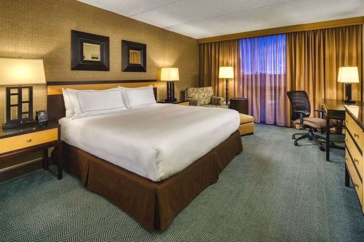 A hotel room with a large bed, two bedside tables with lamps, a desk with a chair, a sofa, and a view of the cityscape through the window.