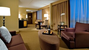 A hotel room with a sofa, armchair, and ottoman. There is a table with a bottle and candles. The room has a view of the city through large windows. - DoubleTree by Hilton Chicago - Arlington Heights (Arlington Heights)