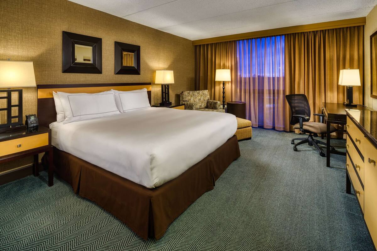 A hotel room with a large bed, two bedside tables with lamps, a desk with a chair, a small sofa, and a view of the city through the window.