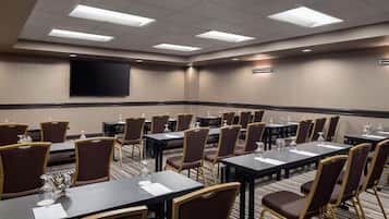 A conference room with multiple tables and chairs, a flat-screen TV on the wall, and a carpeted floor.