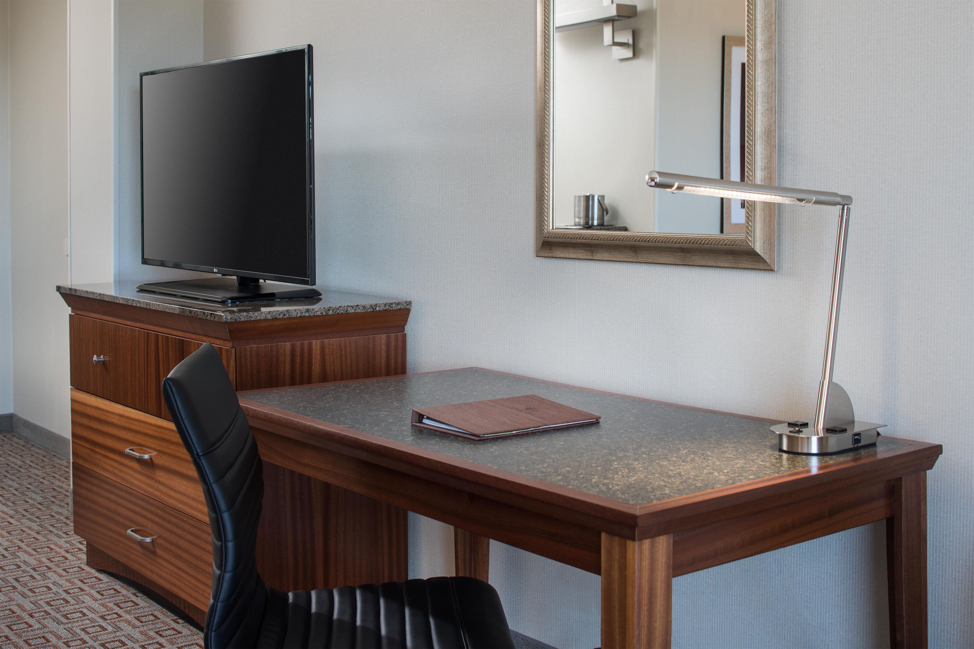 A hotel room with a wooden desk, a television, a mirror, and a lamp.
