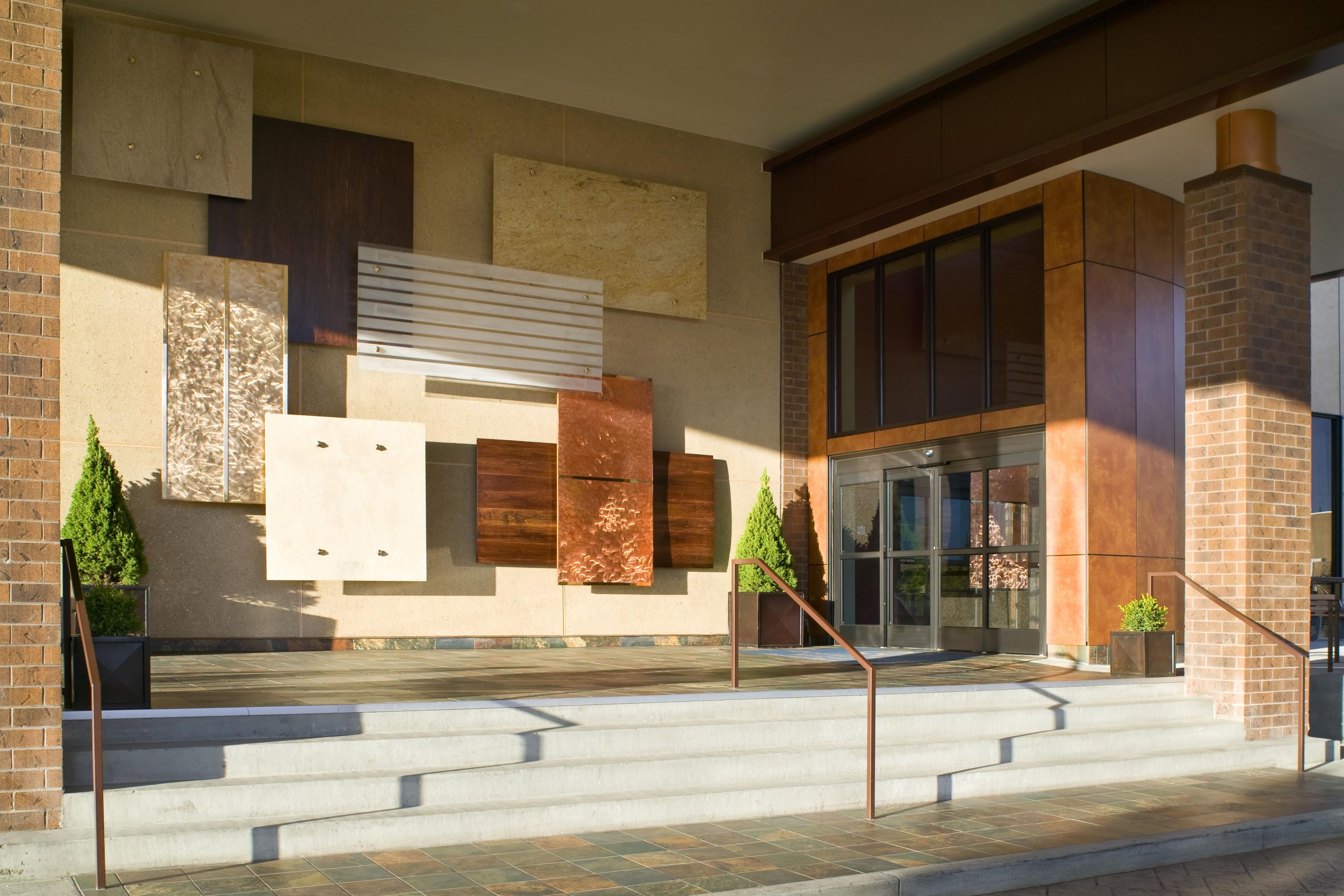 A building entrance with a set of stairs leading up to a glass door, a brick wall with decorative panels, and a metal railing.