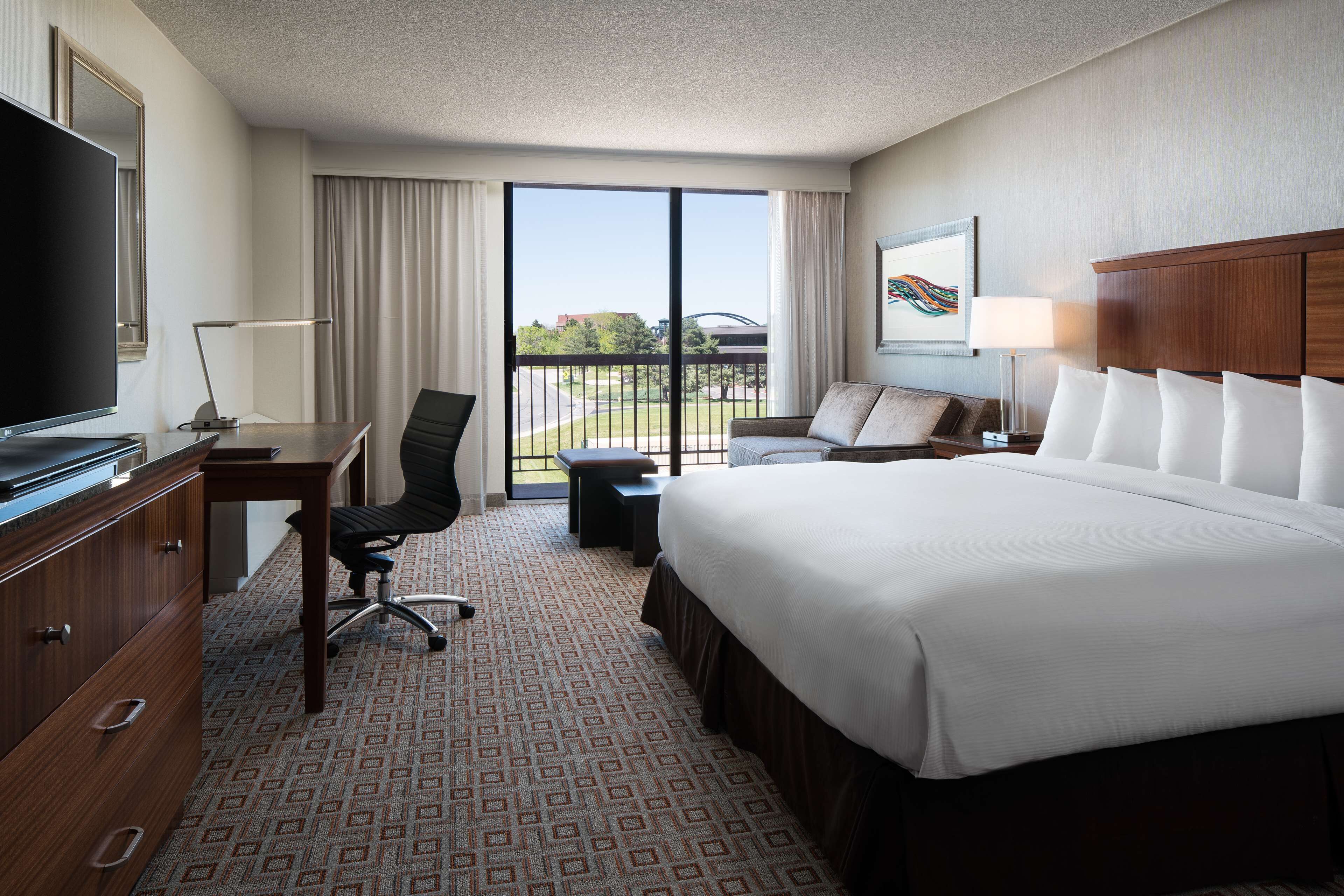A hotel room with a large bed, a desk with a computer, a TV, a chair, and a balcony with a view.