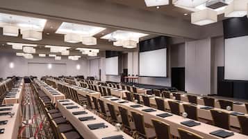 A conference room with long tables, chairs, a projector screen, and ceiling lights.