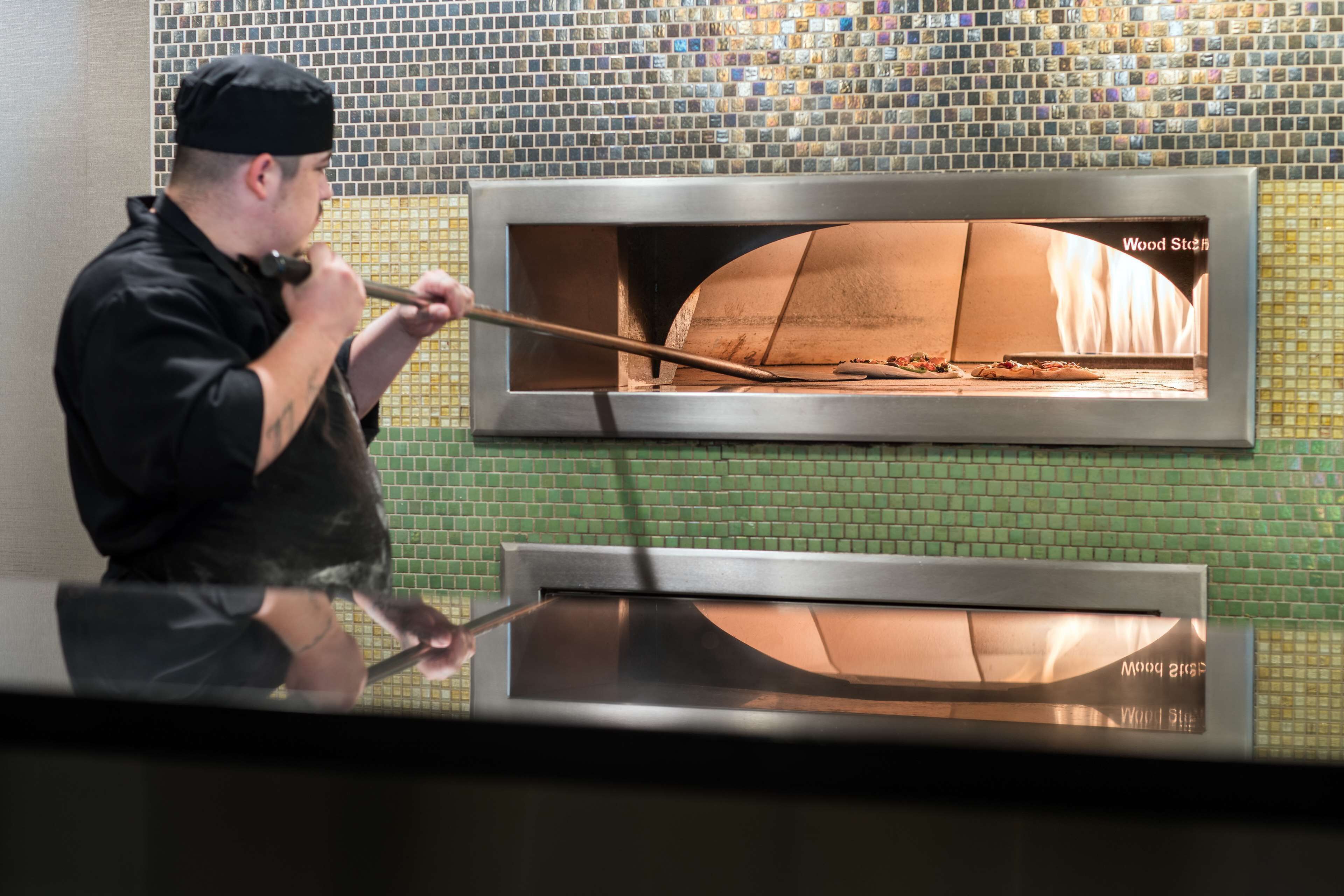 Une personne utilise un outil à long manche pour manipuler un four à pizza portant l’inscription « Wood Stone », le tout se détachant sur un mur recouvert de carreaux verts.