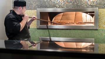 Une personne utilise un outil à long manche pour manipuler un four à pizza portant l’inscription « Wood Stone », le tout se détachant sur un mur recouvert de carreaux verts.
