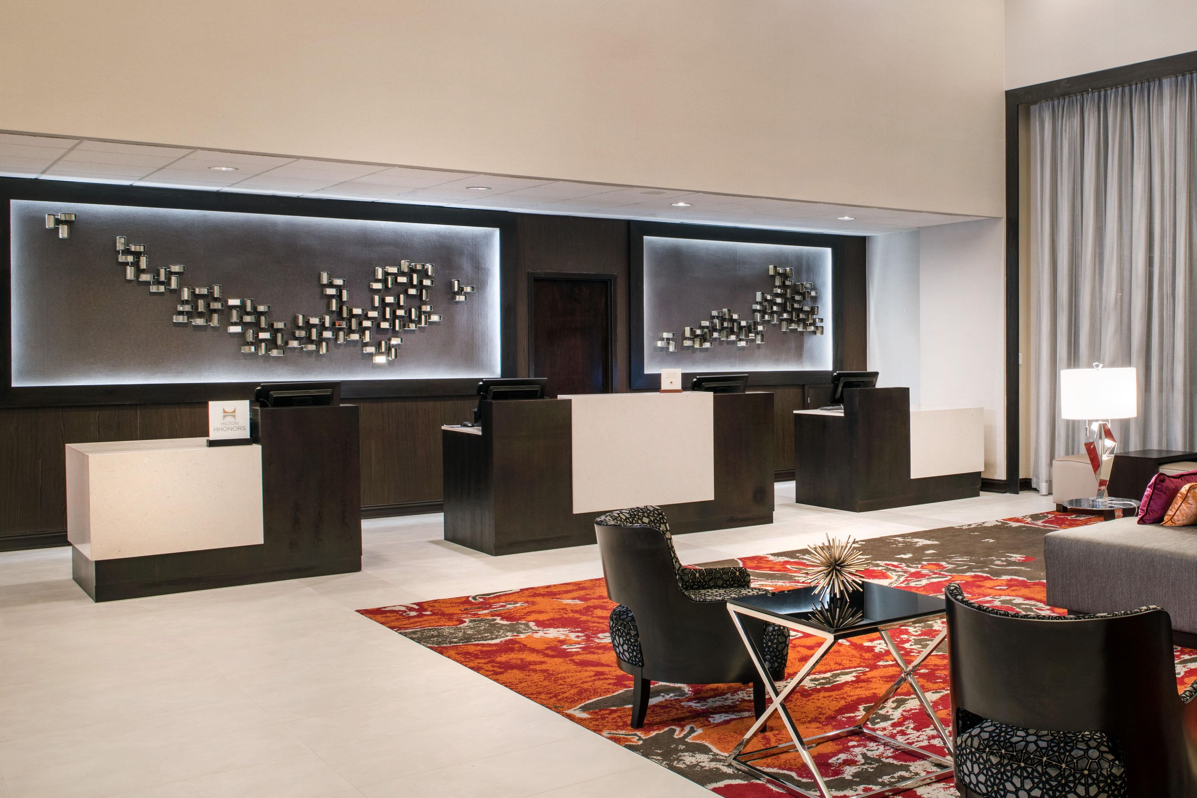Un hall d’hôtel moderne avec deux comptoirs d’accueil, un espace salon avec des fauteuils, un tapis à motifs et une grande fenêtre avec des rideaux.
