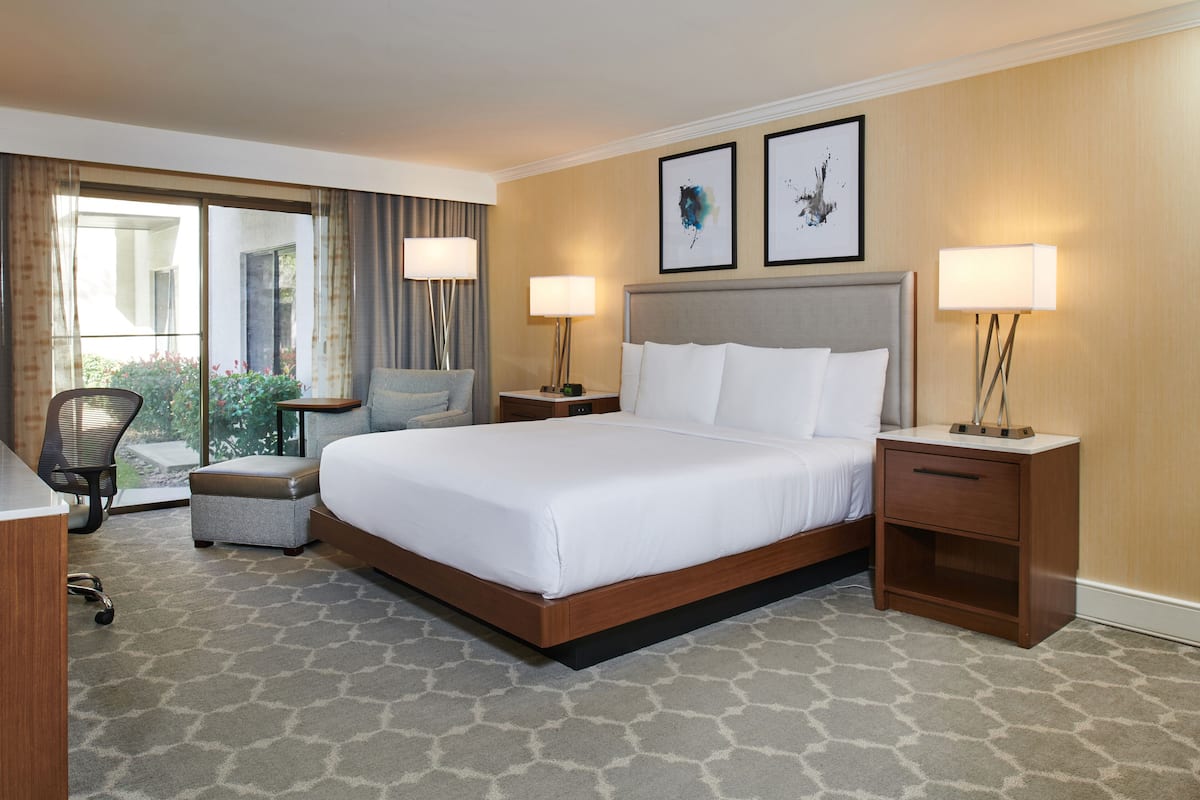 A hotel room with a large bed, two bedside tables with lamps, a chair, a small table, and a view of the outdoors.