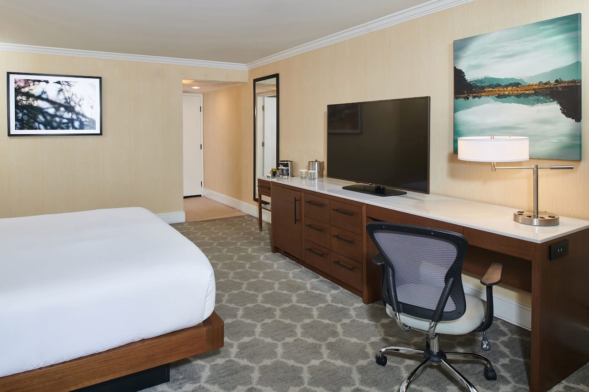 A hotel room with a bed, a desk with a TV, a chair, and a painting on the wall.