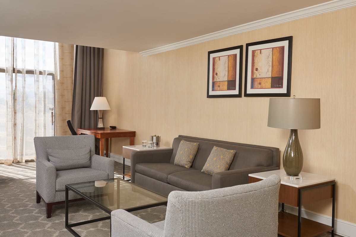 A modern hotel room with a grey sofa, two armchairs, a glass coffee table, a side table with a lamp, and artwork on the wall.