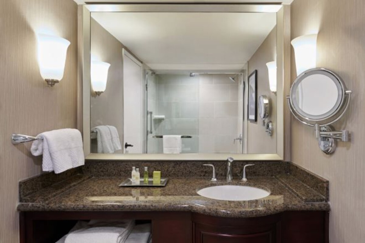 A hotel bathroom with a granite countertop, a sink, a mirror, and two wall-mounted light fixtures.