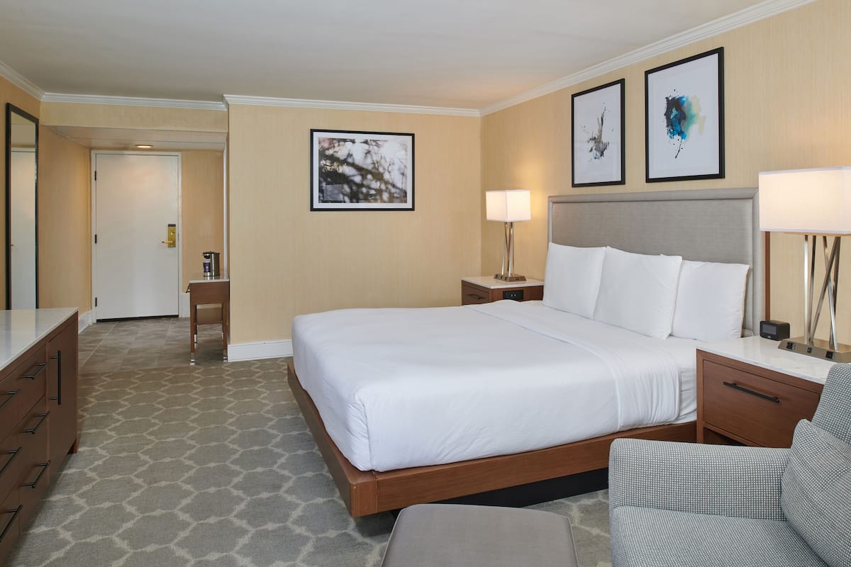 A hotel room with a large bed, a desk, a chair, and a nightstand with a lamp.