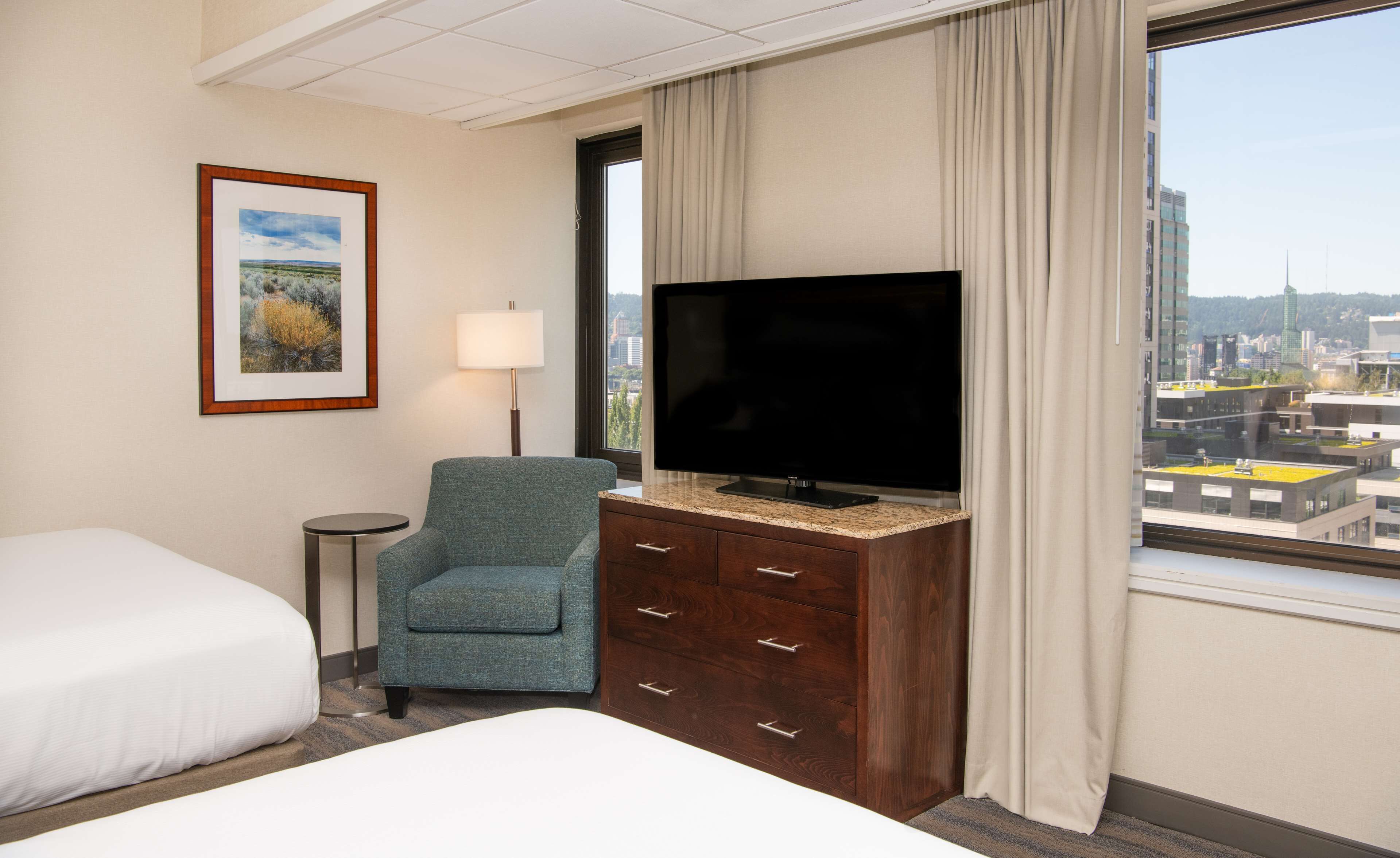 Une chambre d’hôtel avec un lit, une chaise, un téléviseur posé sur une commode en bois, une lampe et une fenêtre donnant sur la ville.