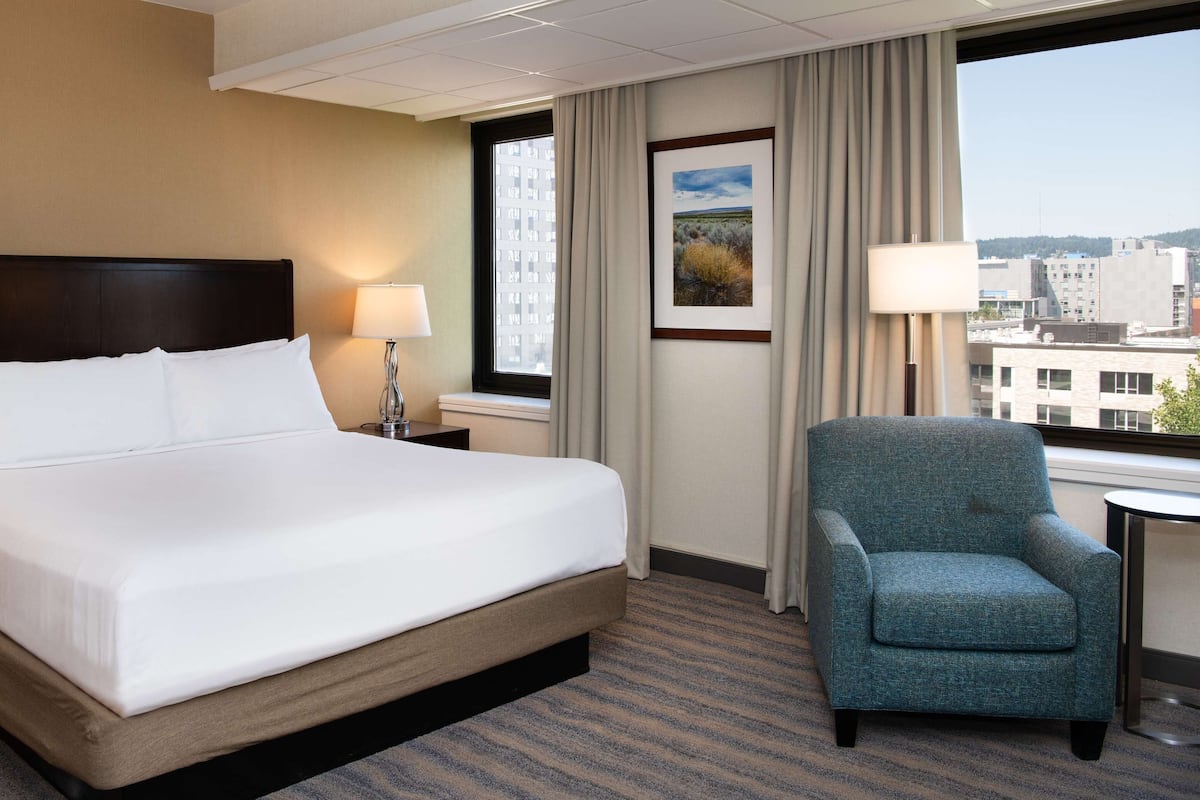 A hotel room with a large bed, two bedside lamps, a chair, and a window with a city view.