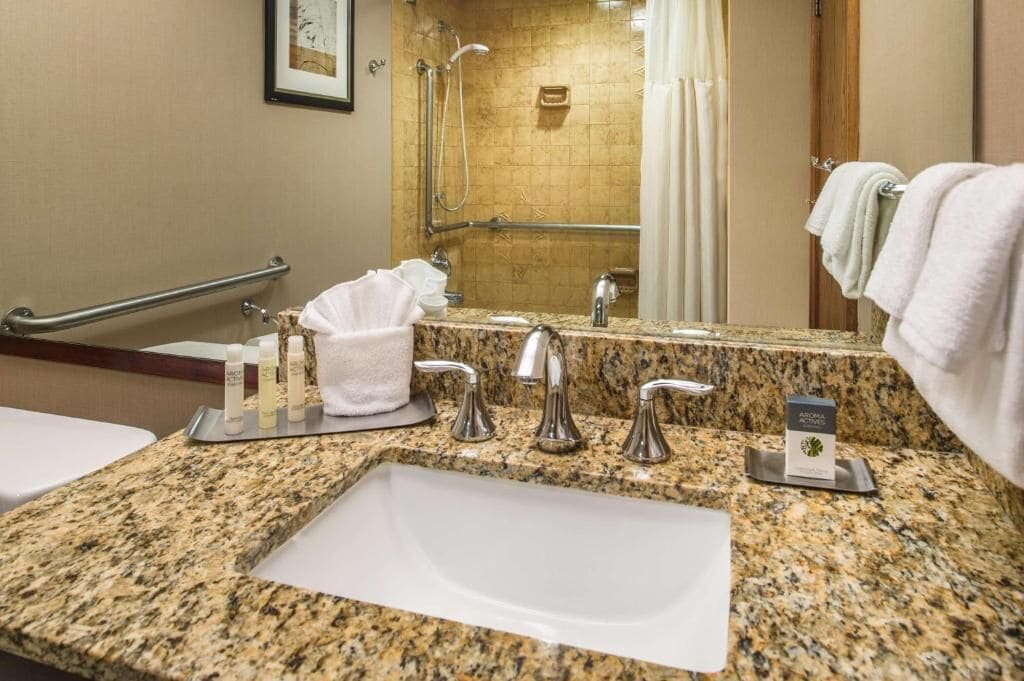 A bathroom with a granite countertop, a sink, a mirror, a shower with a handrail, and a towel rack.