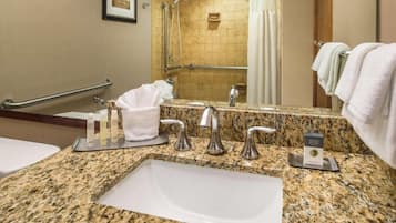 A bathroom with a granite countertop, a sink, a mirror, a shower with a handrail, and a towel rack.