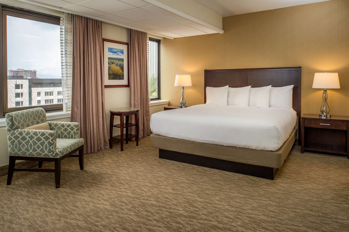 A hotel room with a large bed, two bedside tables with lamps, a chair, a small table, and a view of buildings through a window.