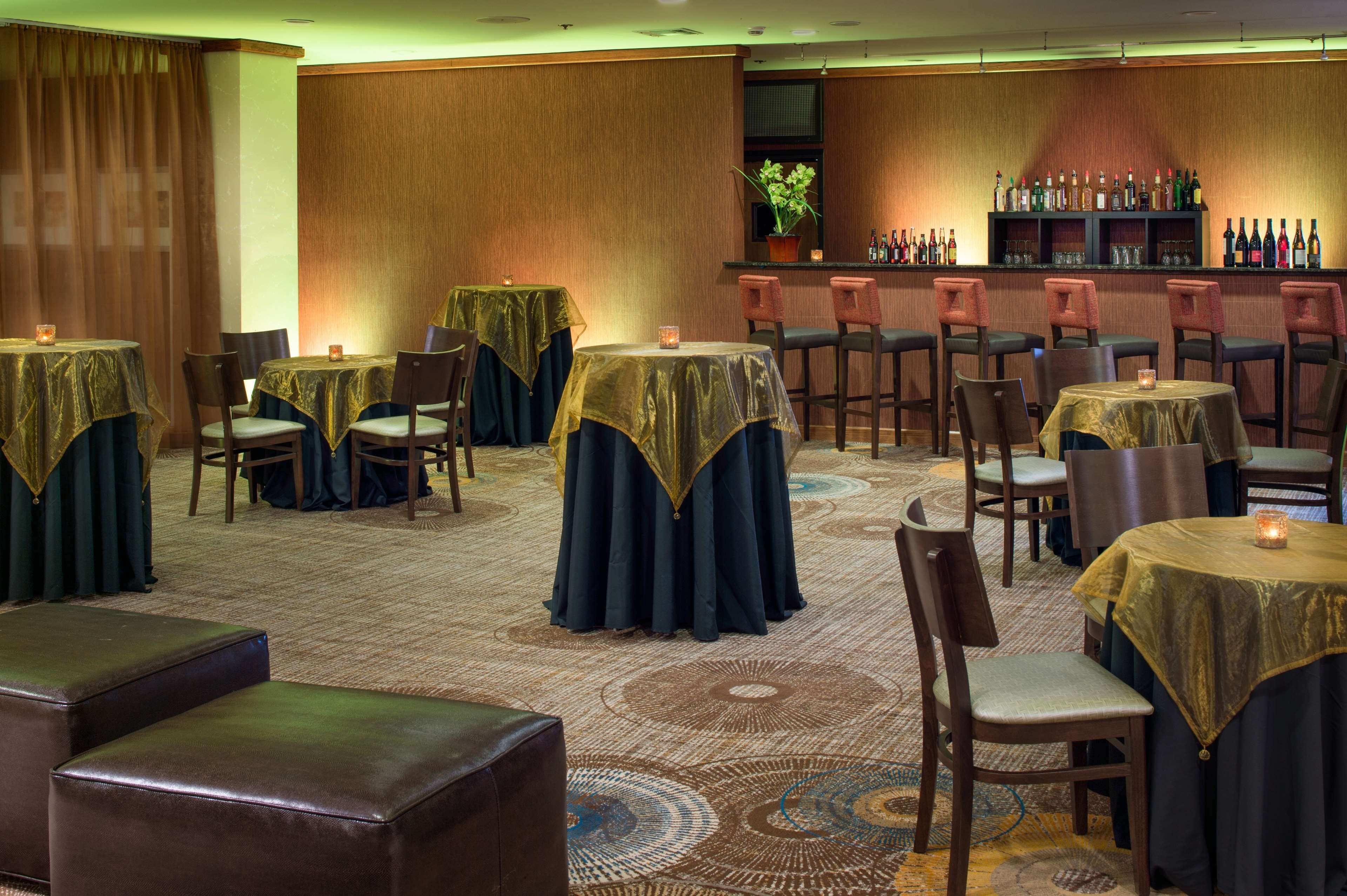 A hotel dining area with round tables covered with gold tablecloths, wooden chairs, and a bar area with shelves of bottles.