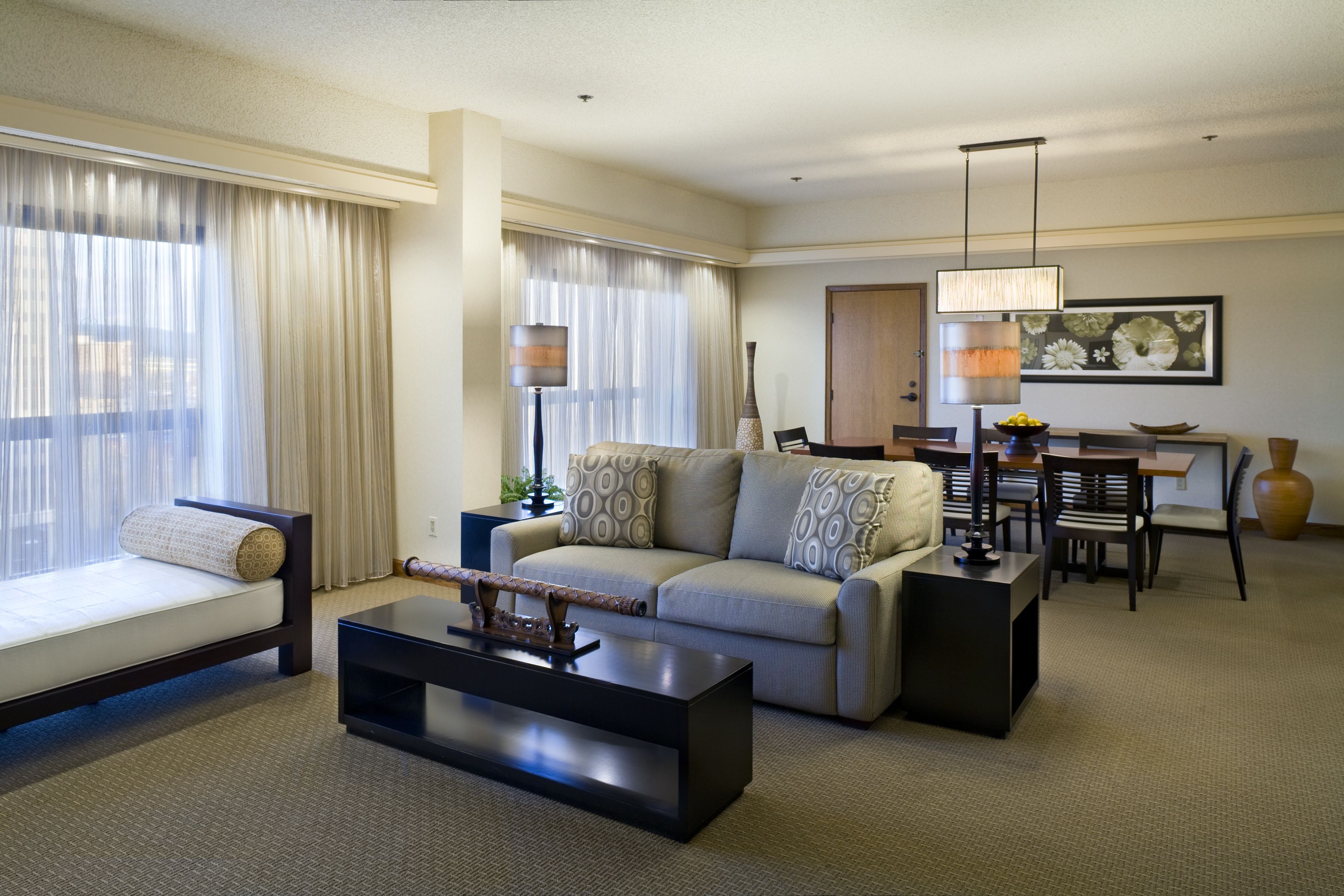 Habitación de hotel con sofá, una mesa de centro, una mesa de comedor con sillas y un ventanal con cortinas.
