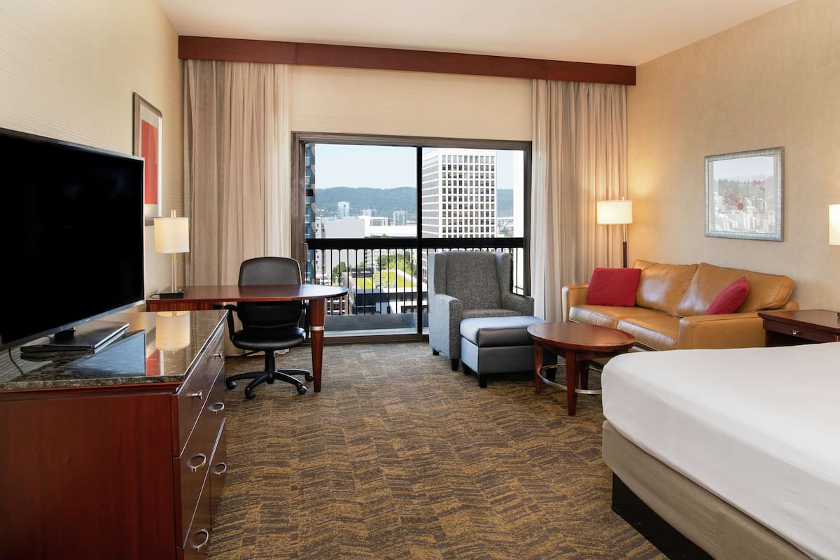 A hotel room with a bed, a desk with a chair, a sofa, and a television. There is a window with a view of buildings and a balcony.