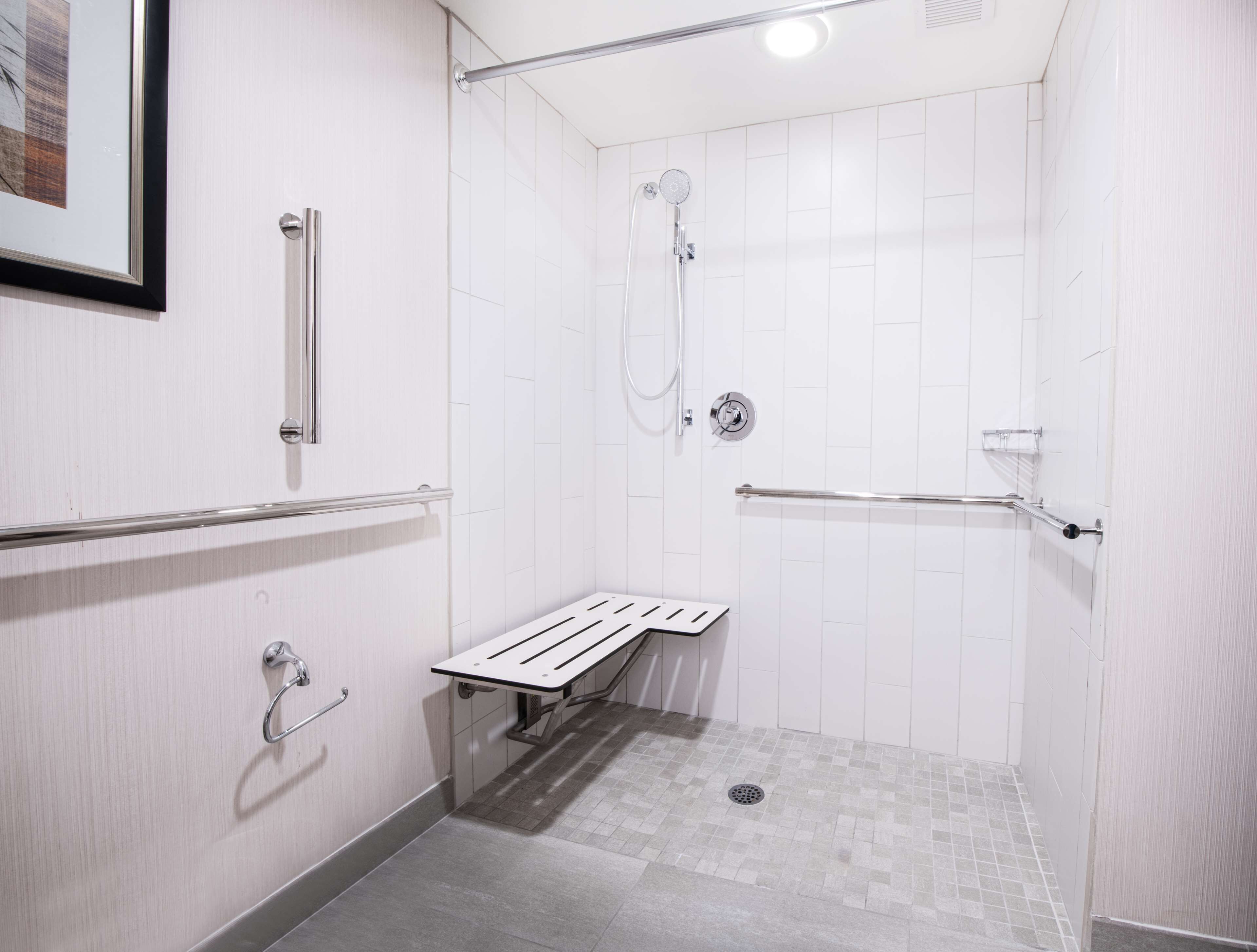 Un área de ducha pequeña y blanca, con azulejos, un banco, barras de apoyo y un cabezal de ducha manual.
