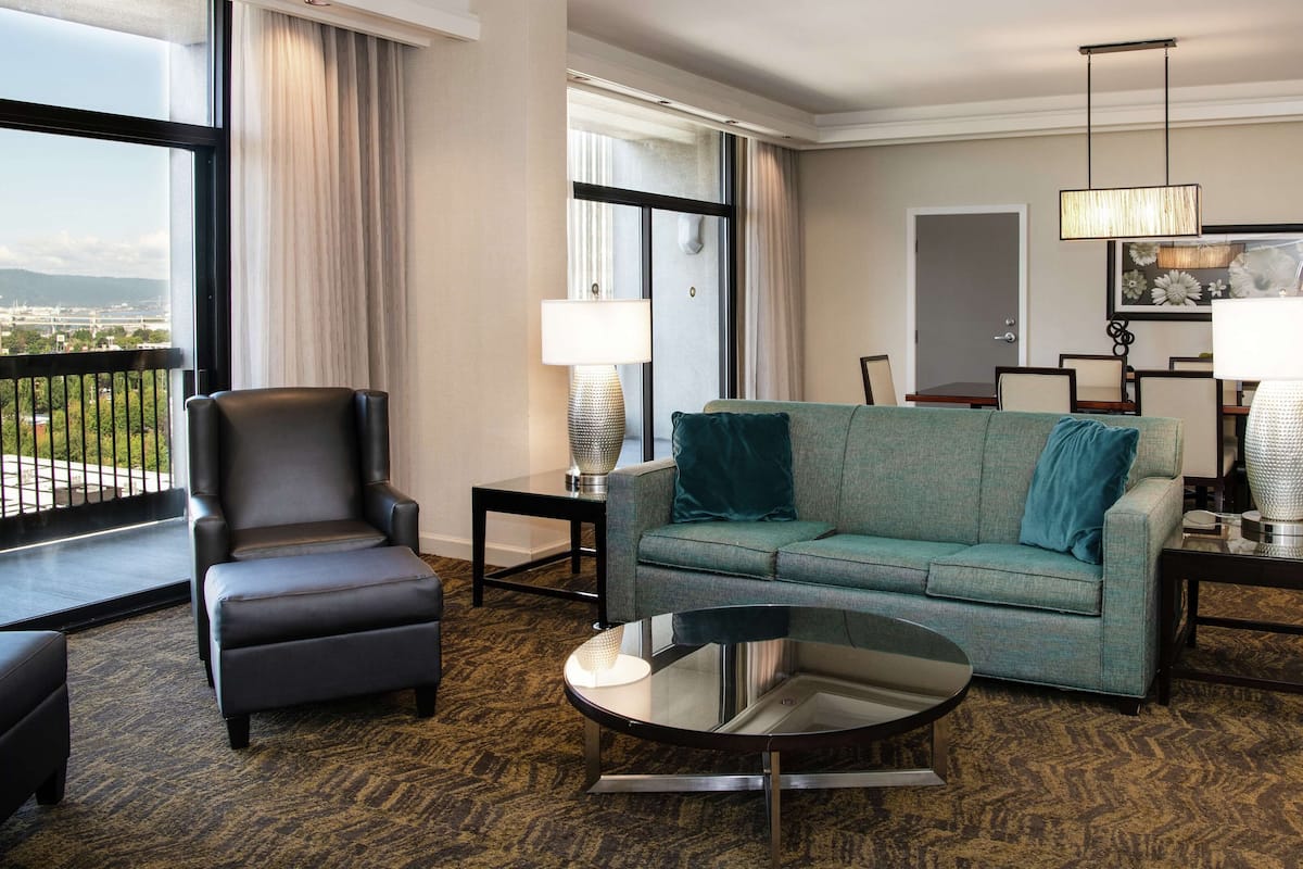 A hotel room with a leather armchair, a teal sofa, a round glass coffee table, and two side tables with lamps. The room has a large window with a view of the city.