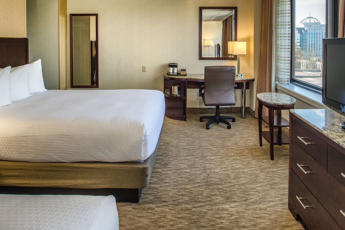 A hotel room with a large bed, a desk with a chair, a television, and a view of the city through the window.