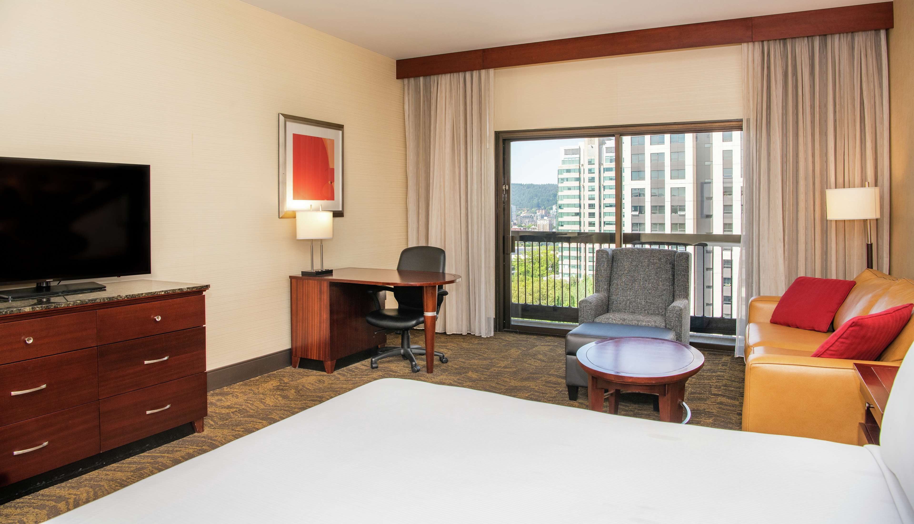 A hotel room with a bed, a desk, a chair, a sofa, a TV, and a window with a city view.