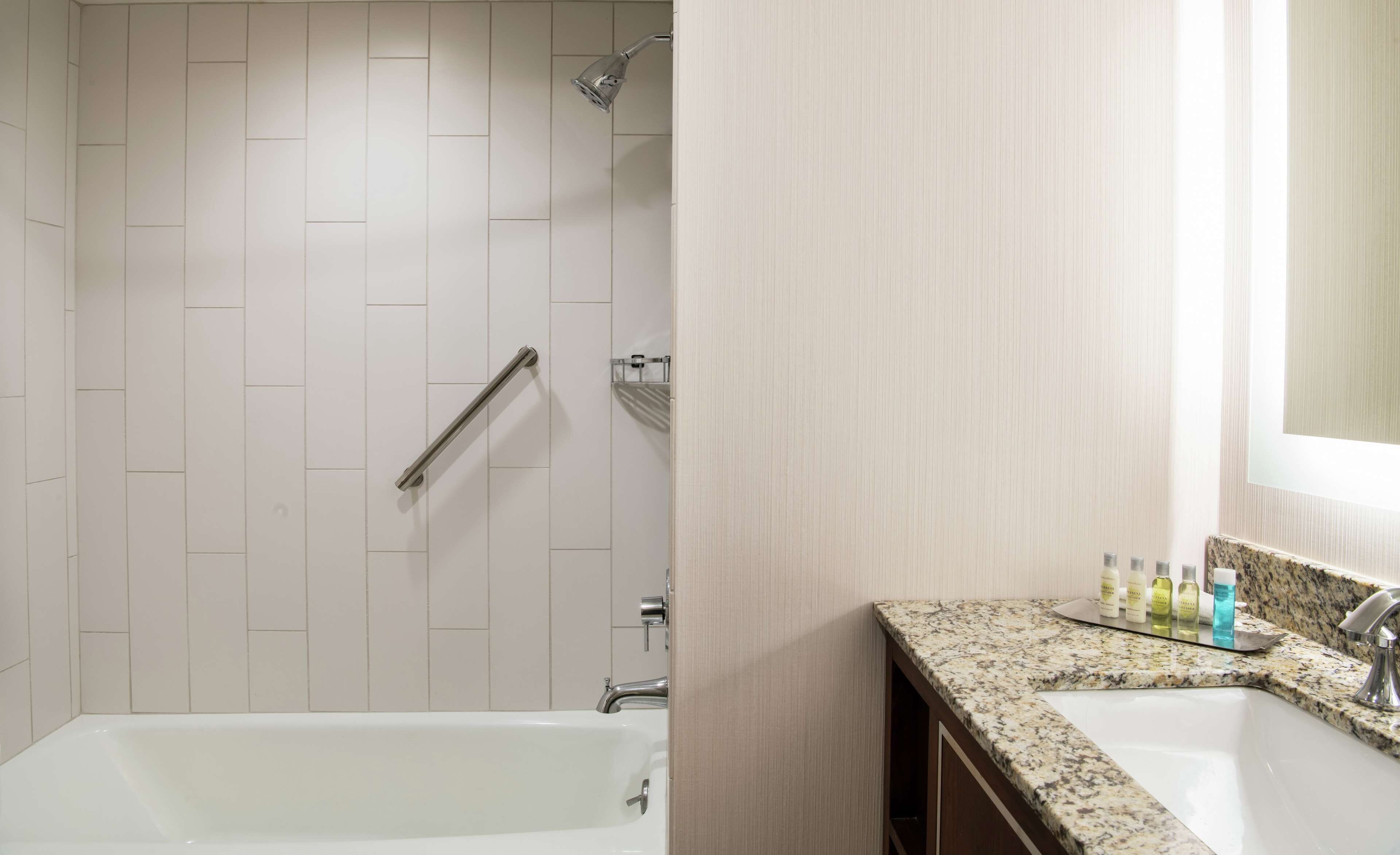 A modern bathroom with a white tiled shower, a grab bar, a bathtub, and a granite countertop with toiletries.