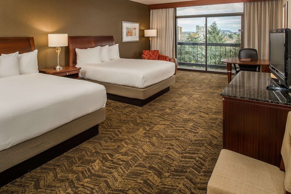 A hotel room with two beds, a desk with a TV, and a large window offering a city view.