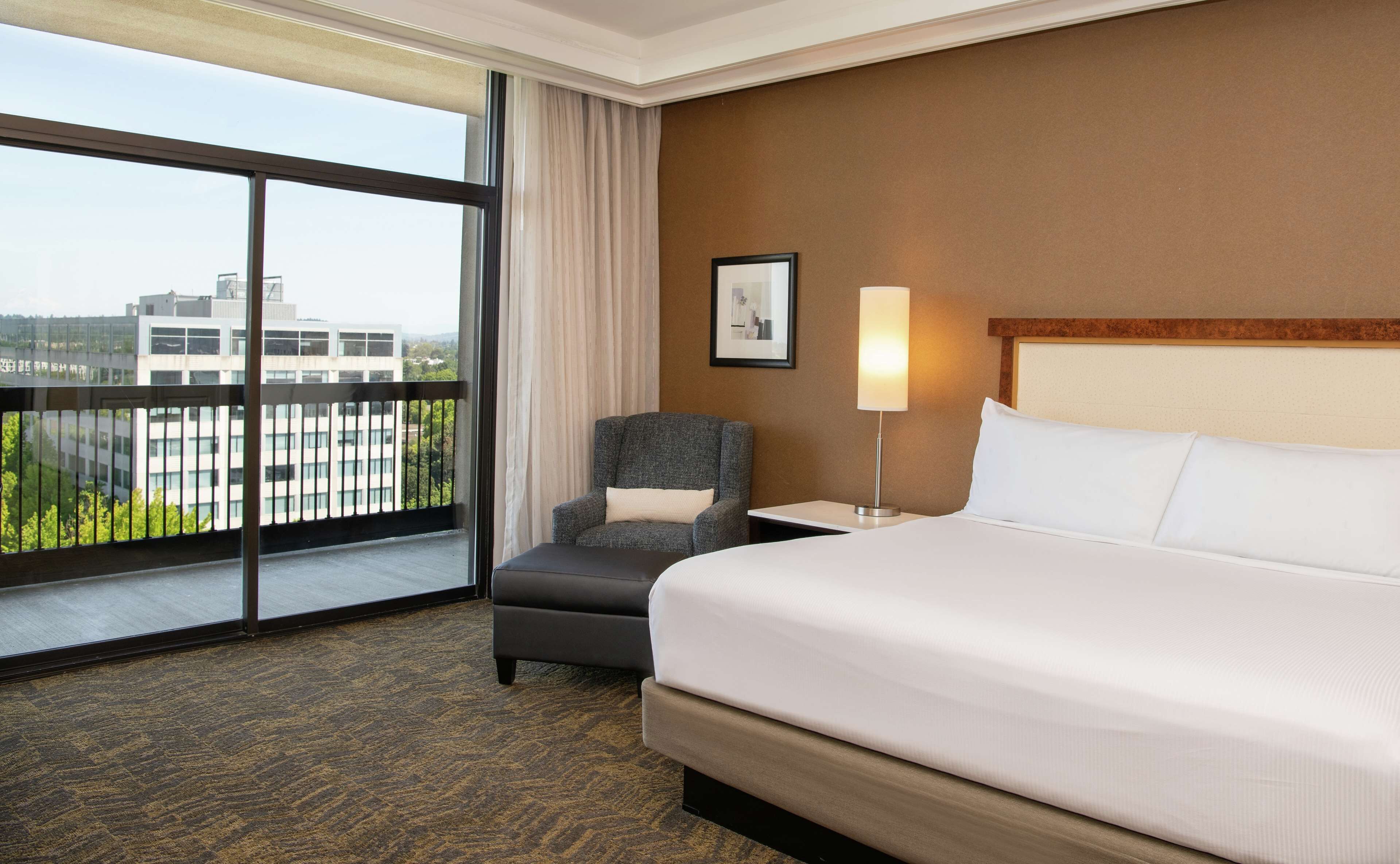 A hotel room with a large bed, a chair, a nightstand, a lamp, and a view of buildings through a large window.