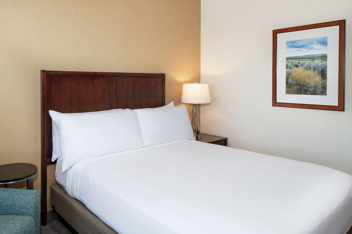 A hotel room with a bed, a nightstand with a lamp, a small round table, and a framed picture on the wall.