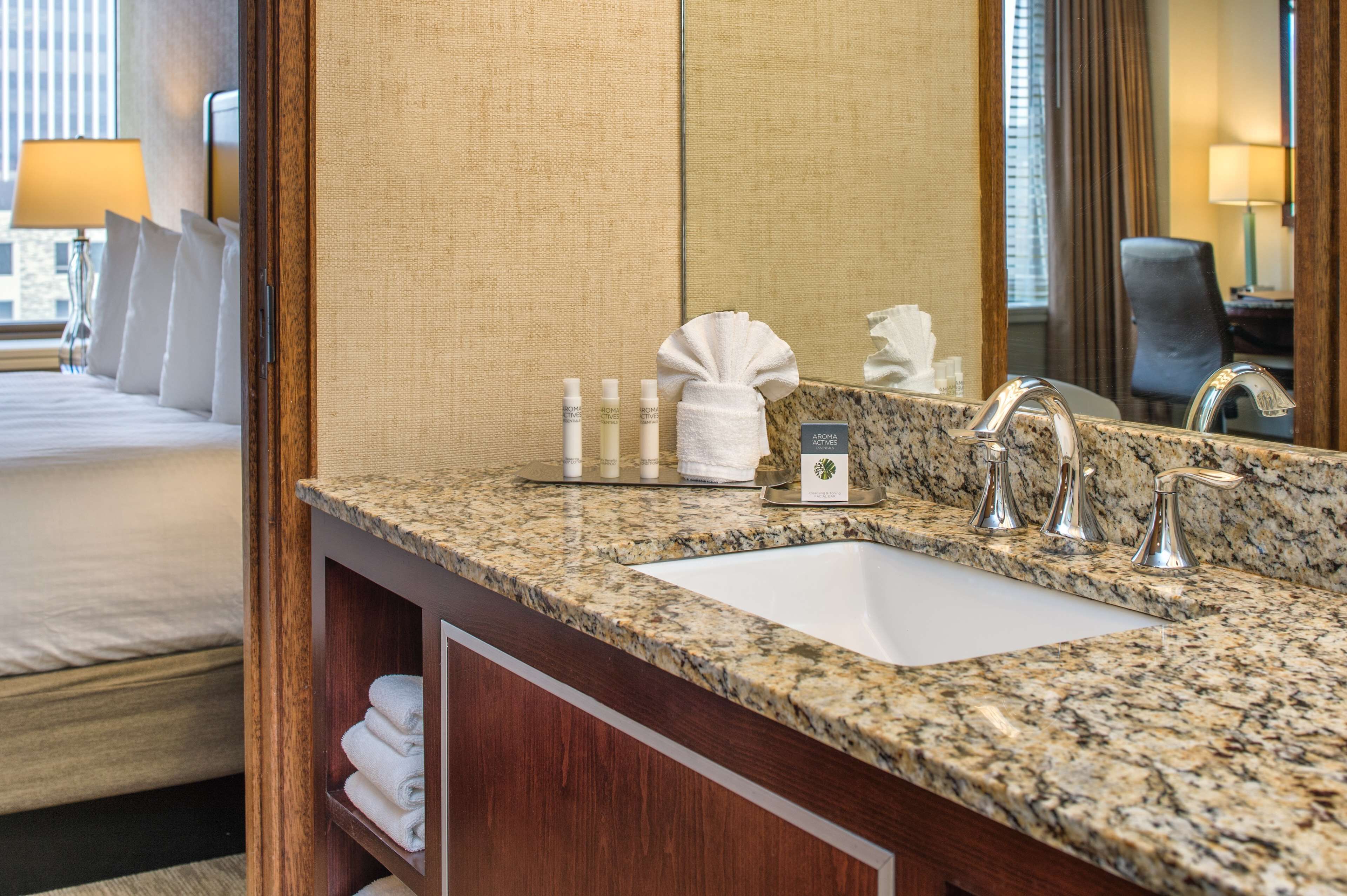 A hotel bathroom with a granite countertop, a sink, and a mirror. There are toiletries on the counter and a towel hanging on the wall.