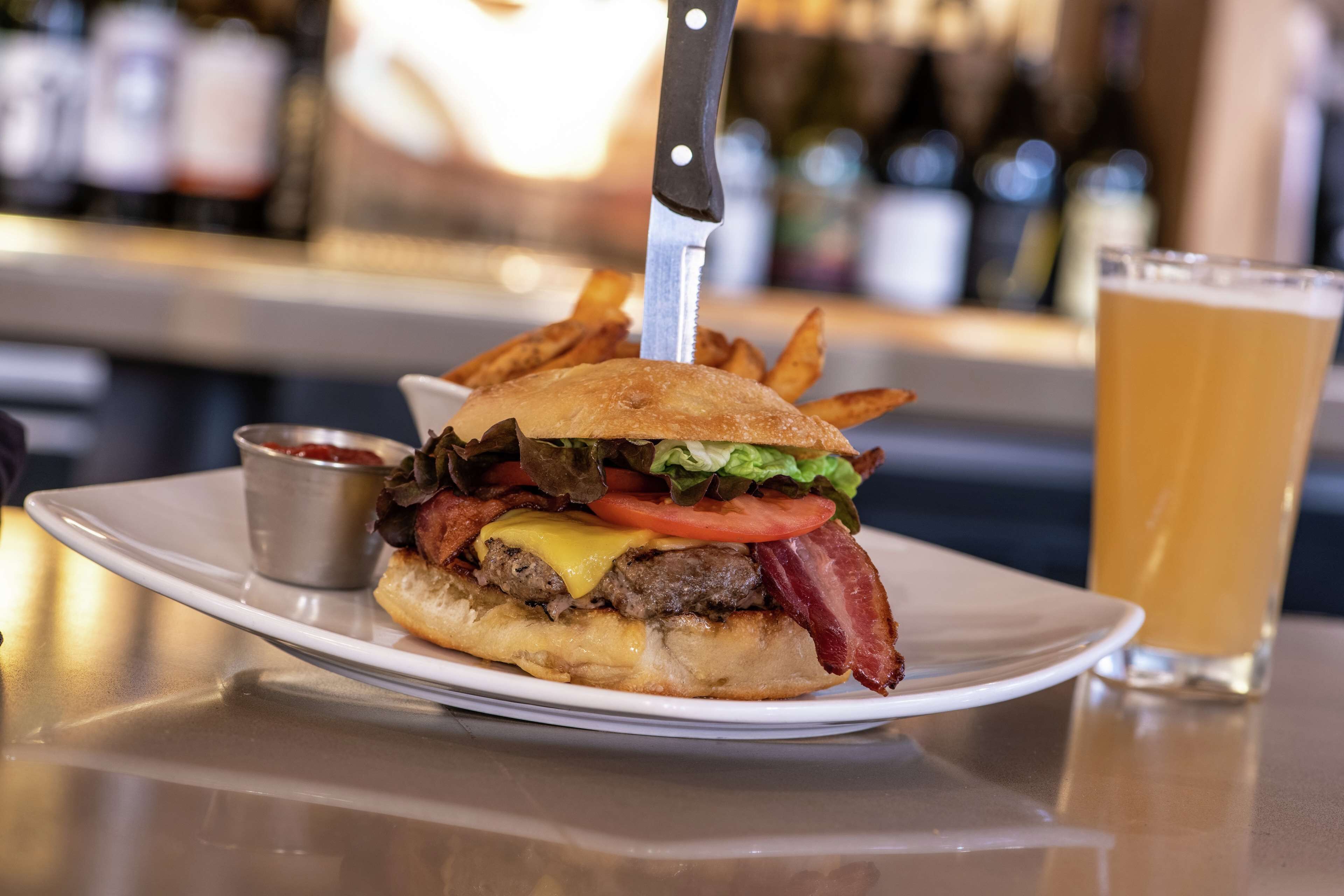 Una hamburguesa con un cuchillo encima, una jarra de cerveza y un recipiente pequeño con salsa sobre una mesa.