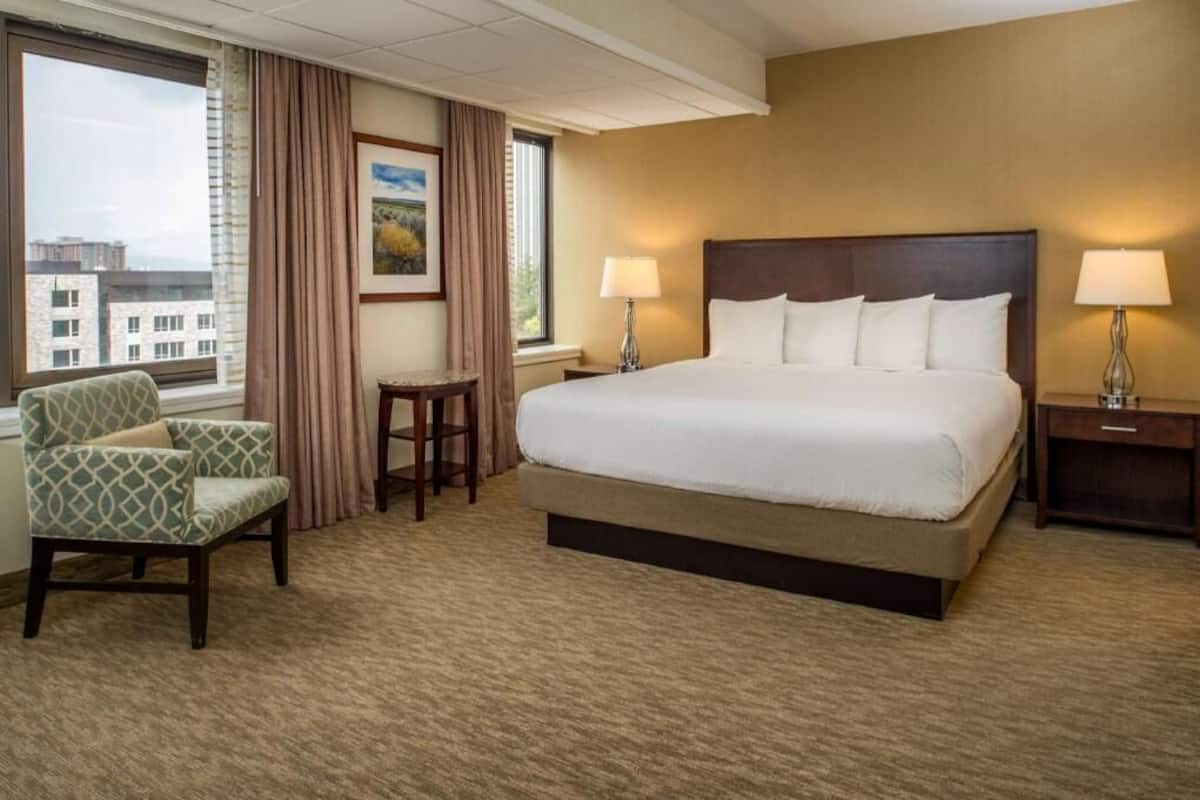 A hotel room with a large bed, two bedside lamps, a chair, a small table, and a view of buildings through the window.