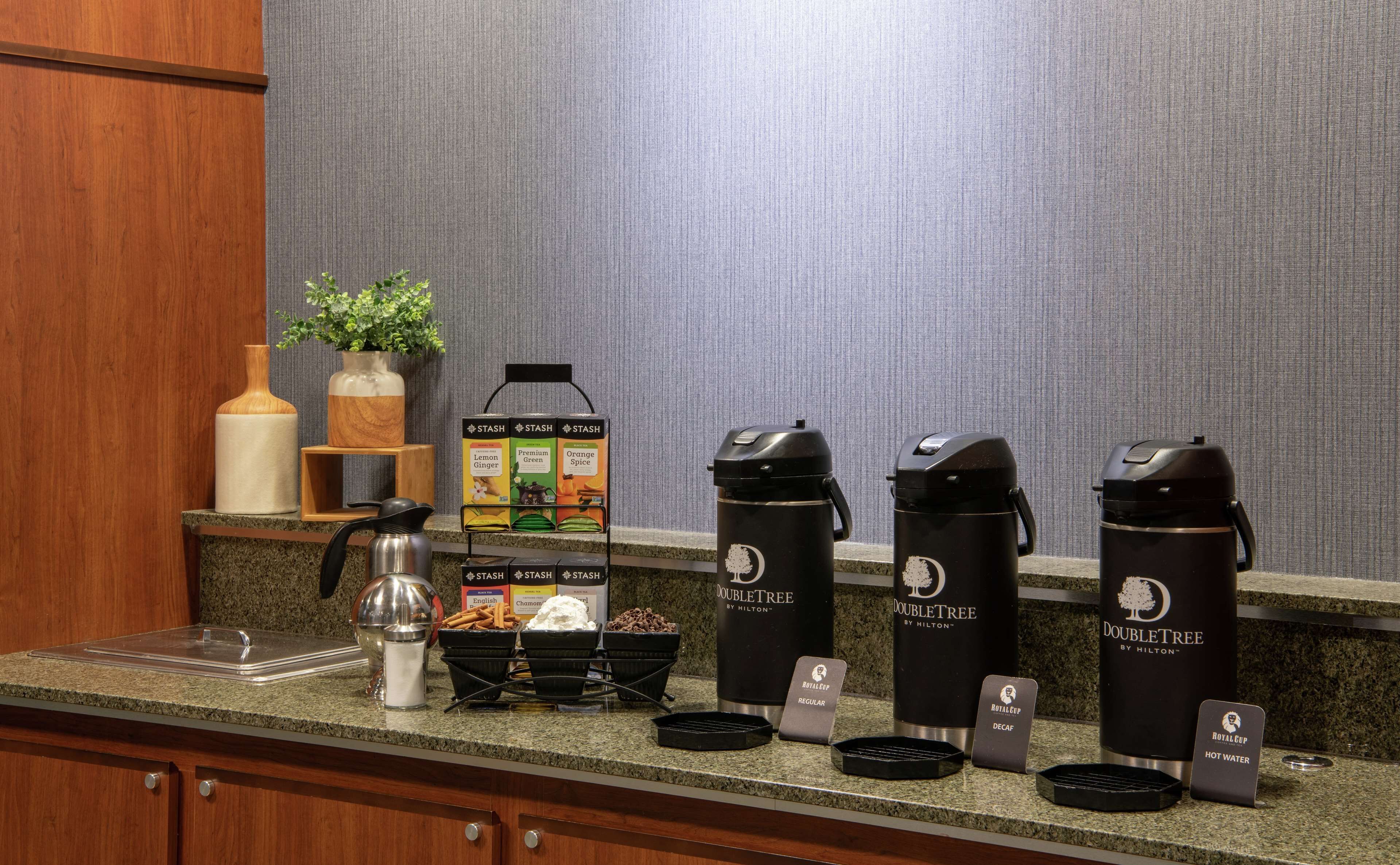 Una estación de café con cuatro máquinas de café DoubleTree, un letrero que indica Royal Cup Regular Decaf, un dispensador de azúcar y una caja de bolsitas de té Stash.