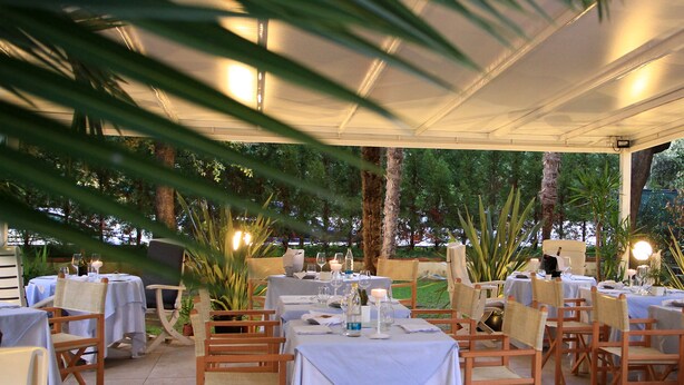 An outdoor dining area with tables set for two, surrounded by chairs and palm trees. - Raffaelli Park Hotel (Forte dei Marmi)