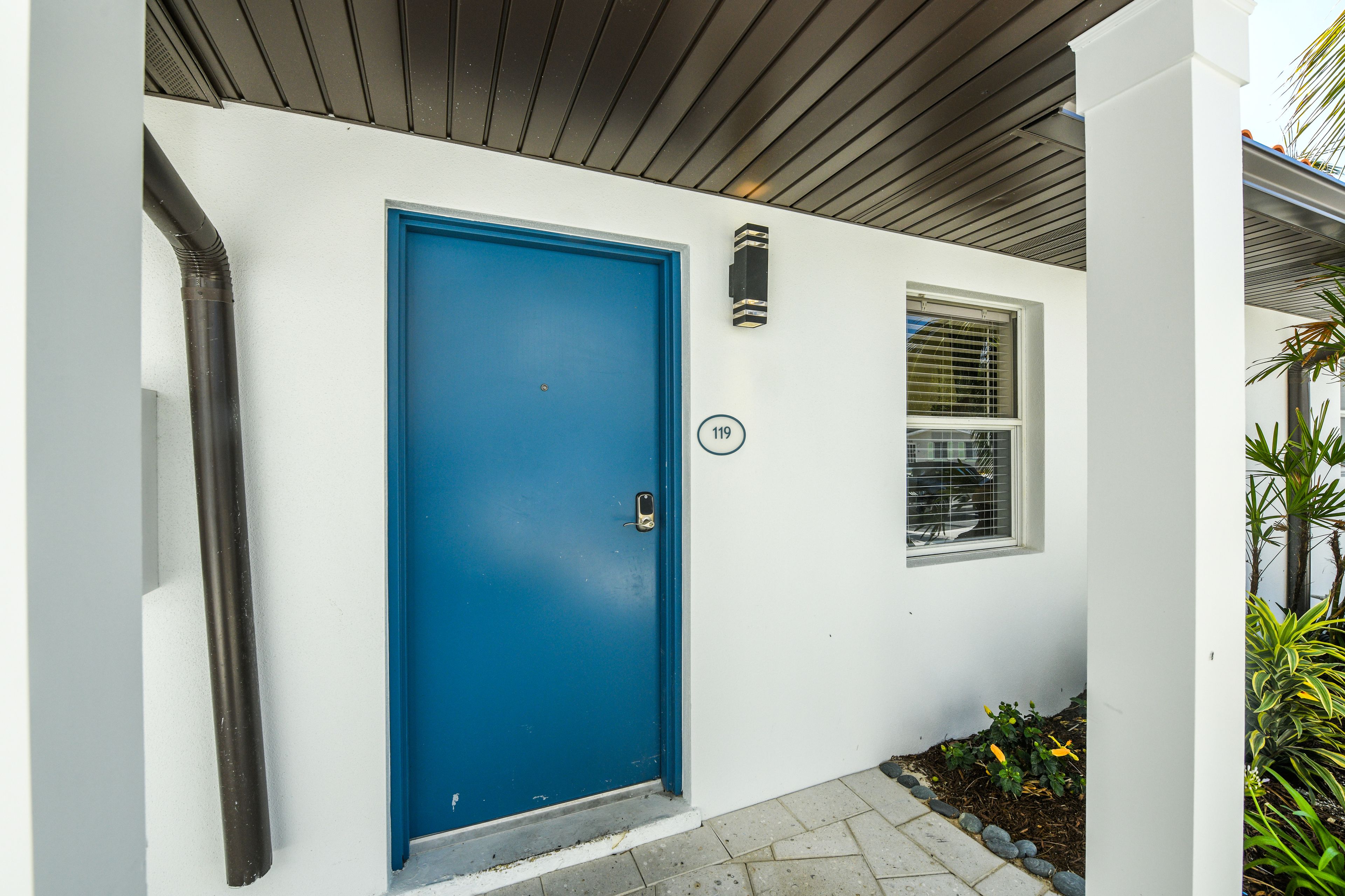 Una puerta azul con un cartel, una ventana con persianas y un pequeño jardín con plantas.