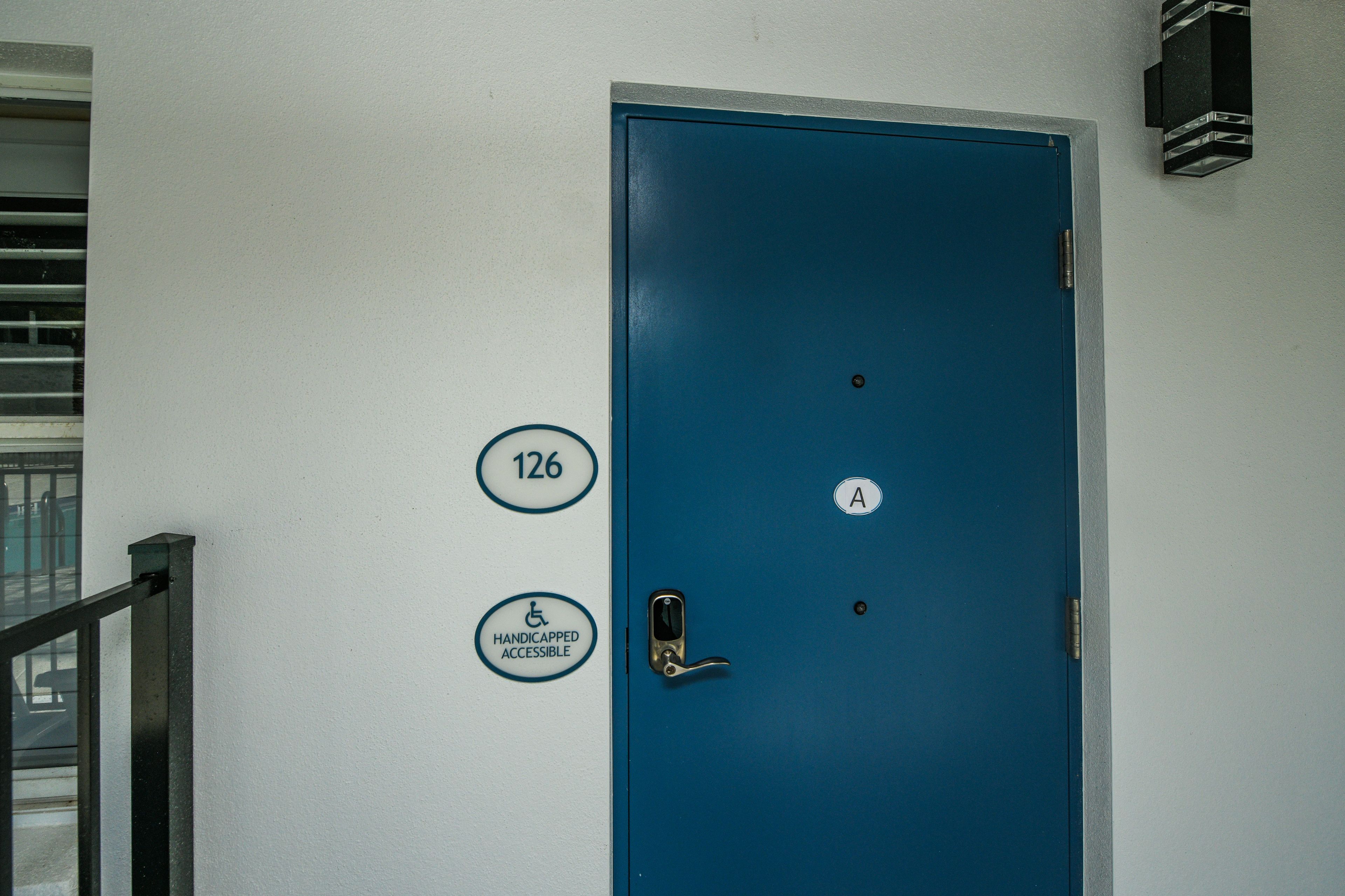 Una puerta azul con señal de accesibilidad para personas con discapacidad, un letrero con el número de la habitación y un letrero con la letra 'A'.