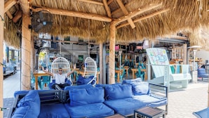Un espace extérieur aménagé avec des canapés et des tables bleus dotés de coussins, un toit de chaume et une enseigne indiquant le « Big Tiko Lounge ».
