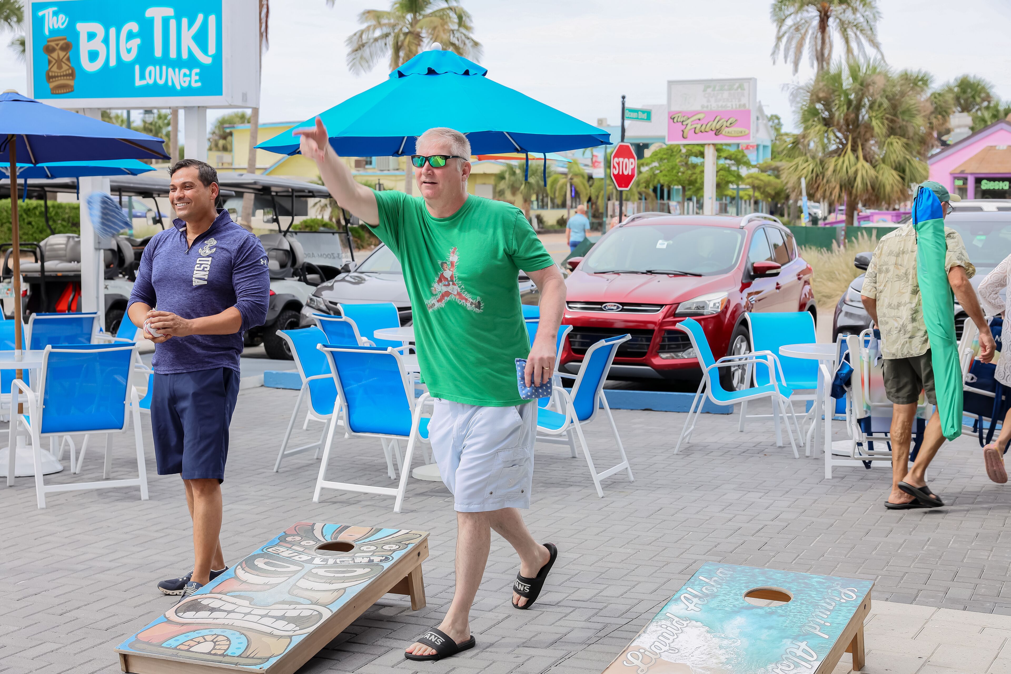 A man walking with a blue umbrella, a sign for "The Big Tiki Lounge," and outdoor seating with blue chairs.