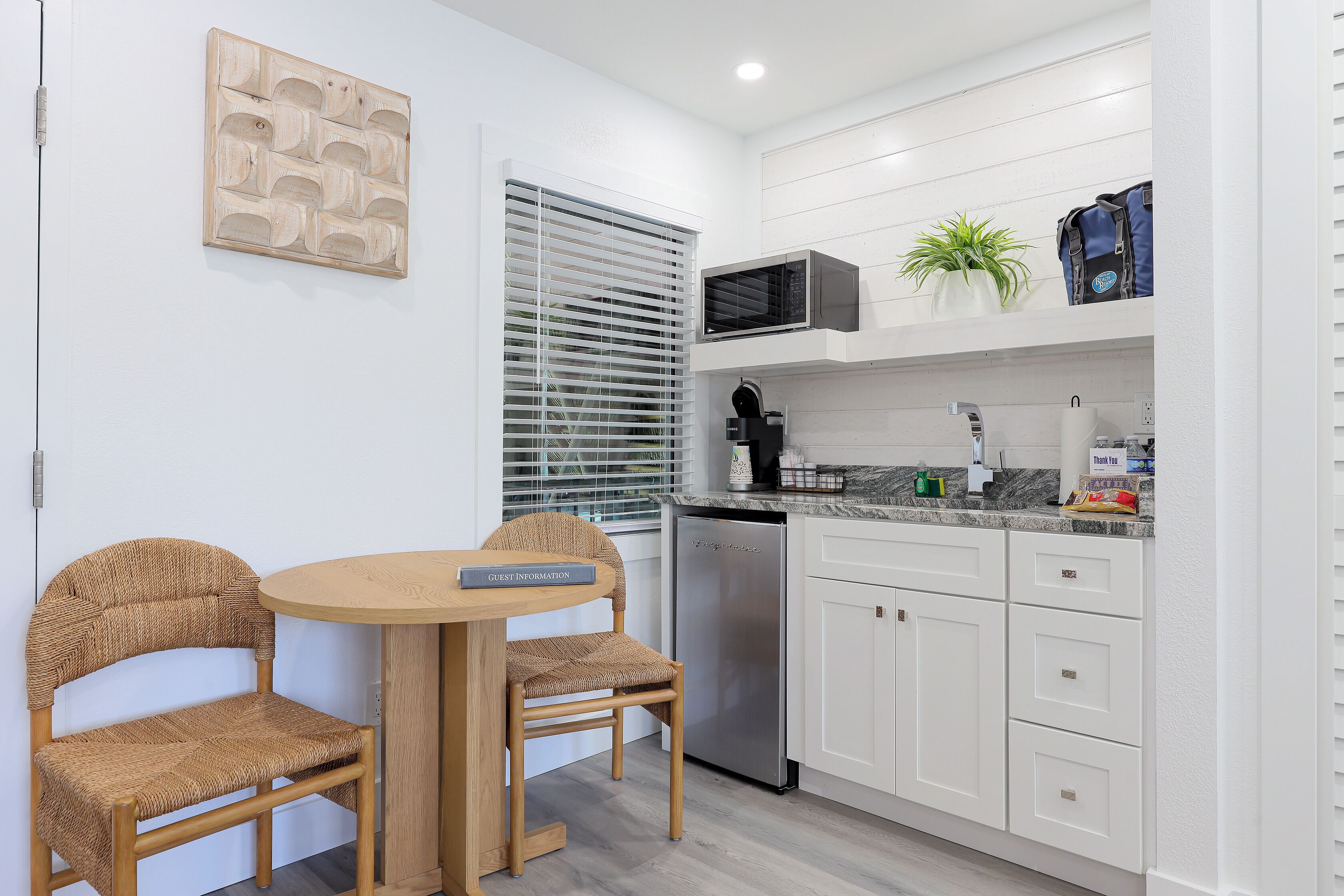 A compact kitchen with white cabinets, a stainless steel dishwasher, and a microwave above the counter. There is a small potted plant on the countertop and a guest information card on the table.