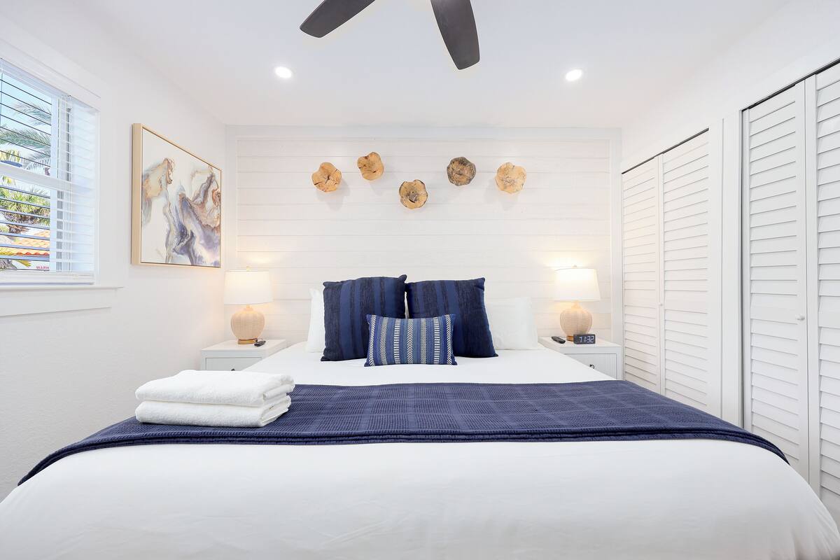 A neatly made bed with a blue blanket, white pillows, and a blue throw. There are two bedside tables with lamps, a wall clock, and a painting above the bed.