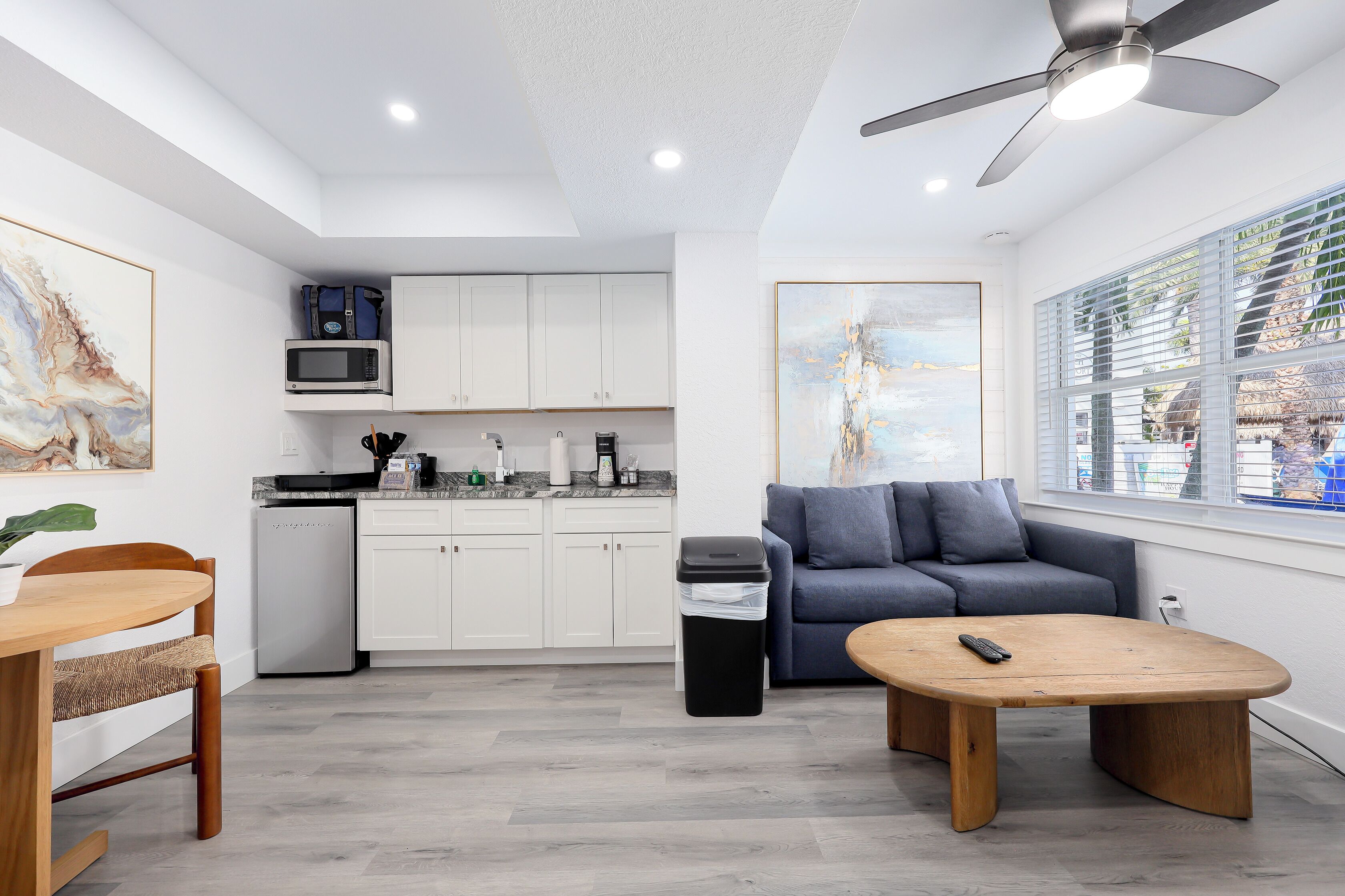Una cocina moderna con armarios blancos, un microondas y una cafetera. Hay un sofá gris y una mesa de madera con un control remoto.
