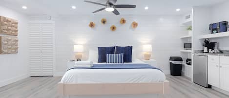 A modern bedroom with a bed, bedside lamps, a ceiling fan, and a kitchen area with appliances.