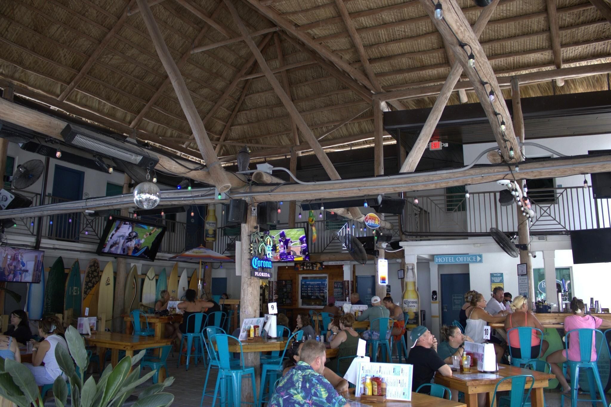 A restaurant with a thatched roof, wooden beams, and surfboards on the wall. There are tables and chairs for dining, and a TV screen displaying a program.