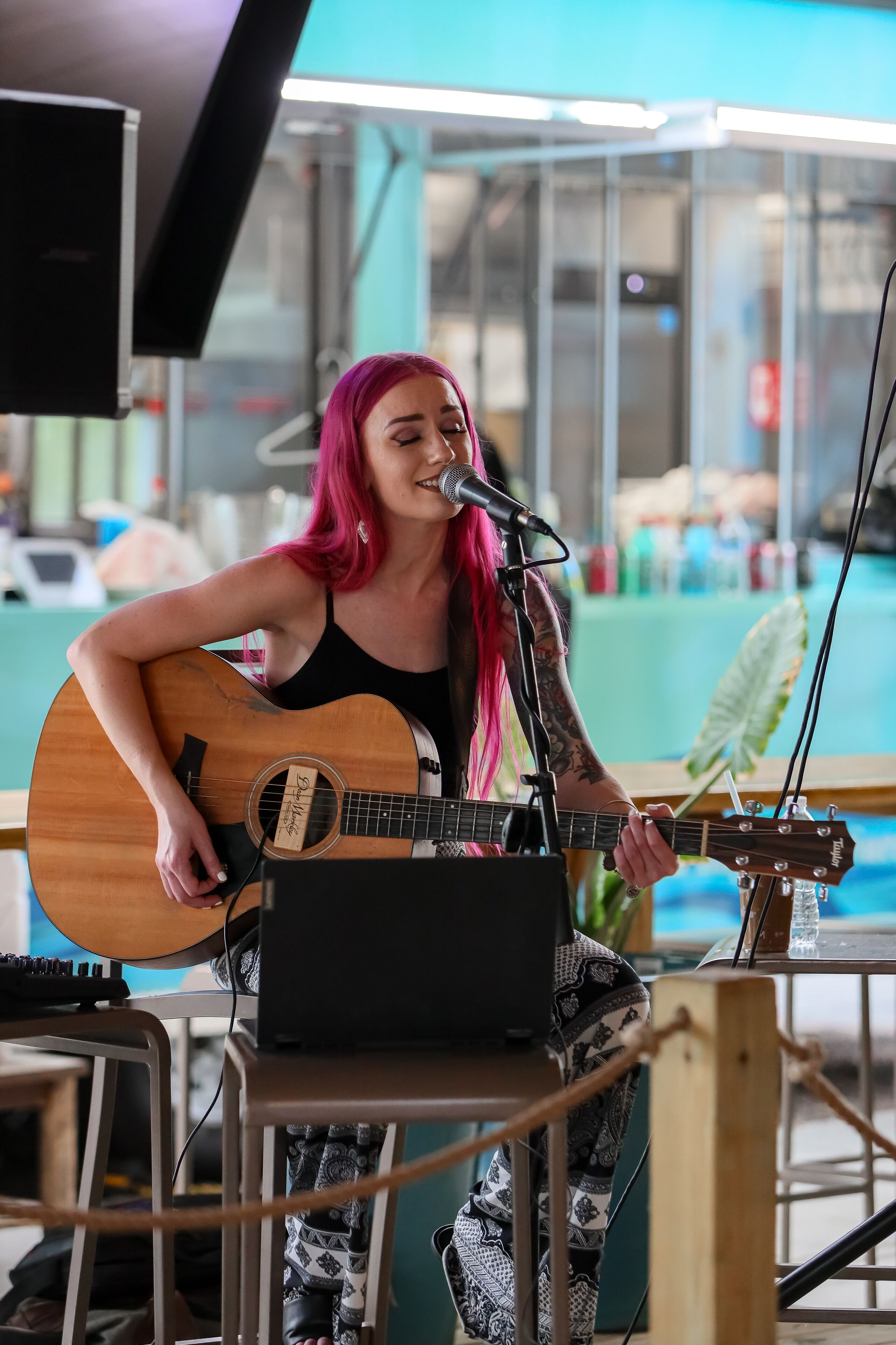 A person playing an acoustic guitar at a microphone stand in a room with a glass partition and a visible outdoor area.