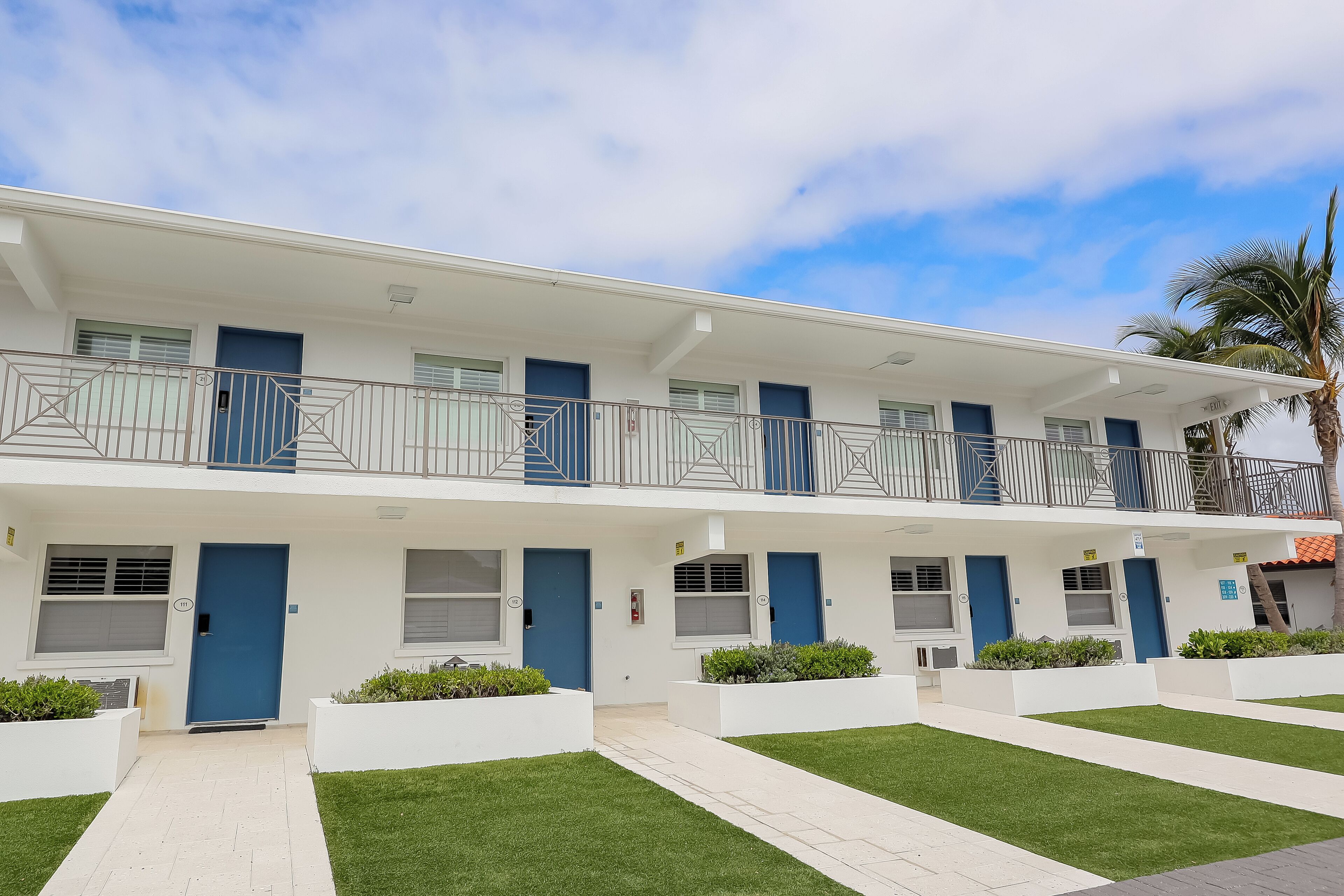 A modern multi-story building with a white facade, blue doors, and a balcony on the upper floor.