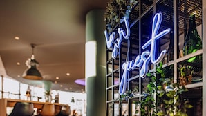 L’intérieur d’un restaurant, avec un panneau néon sur lequel on peut lire « Votre invité » et un autre panneau indiquant « spécialités du chef ».